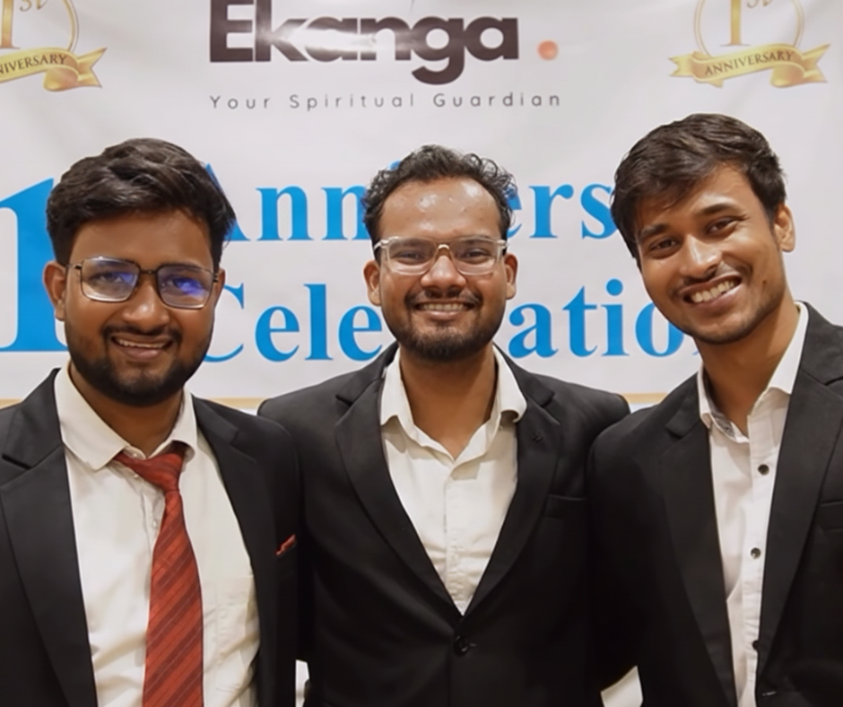 Smiling Ekanga founders in formal suits at Ekanga anniversary celebration event, standing in front of an Ekanga “Your Spiritual Guardian” backdrop.